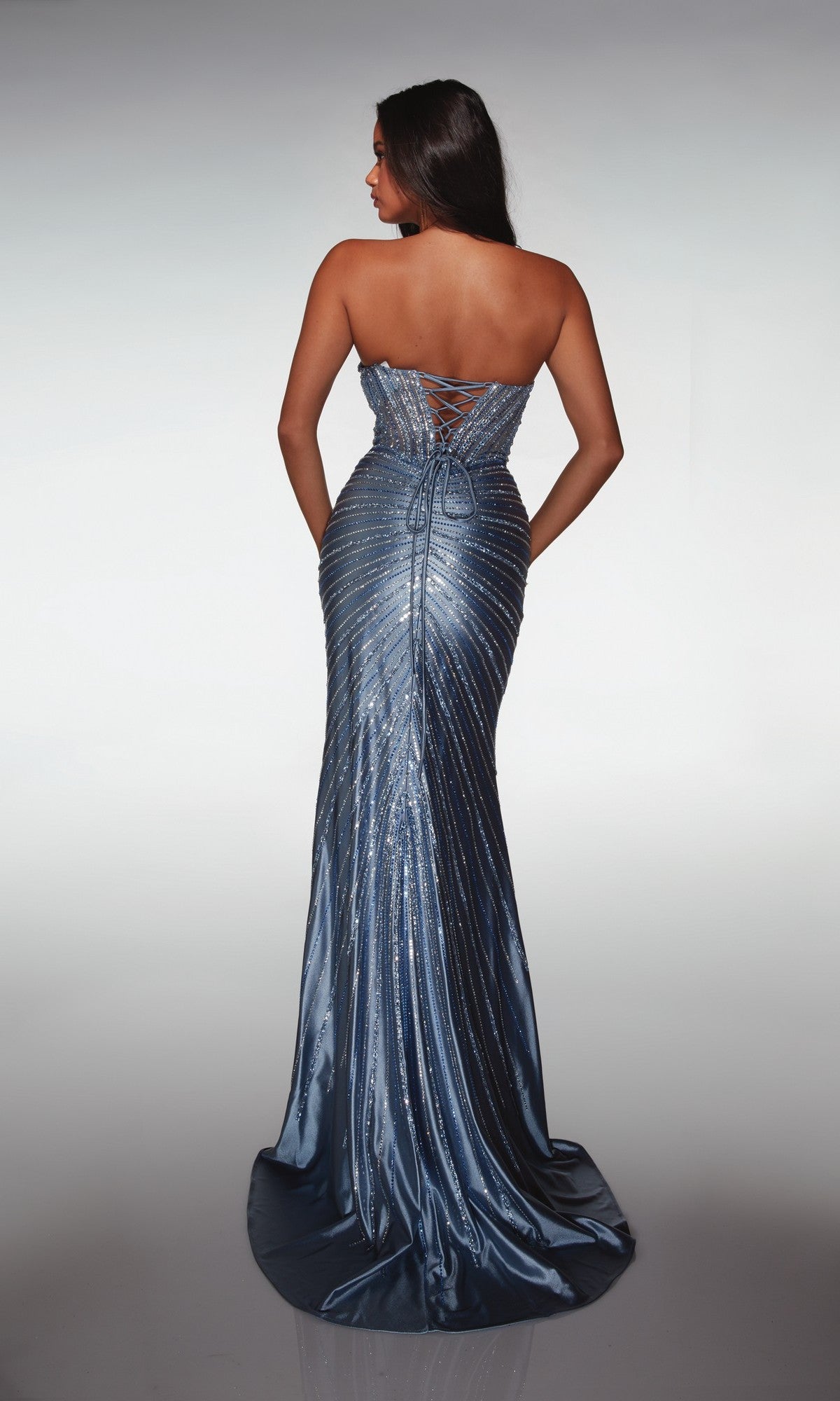A woman models the Alyce 62142 long prom dress—a strapless satin gown with a metallic blue finish, lace-up back, corset bodice detailing, sparkling fitted design, and a slight train—facing away to display its features.
